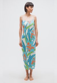 Γυναίκα με αμάνικο, μακρύ φόρεμα μέχρι τον αστράγαλο με μοτίβα από μπλε, πράσινα και πορτοκαλί λουλούδια, όρθια ξυπόλυτη, κοιτάζει προς τα δεξιά σε απλό λευκό φόντο.