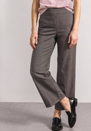 Femme portant un pantalon marron droit à carreaux, un haut rose et des mocassins en cuir noir, debout avec un pied croisé devant l'autre.
