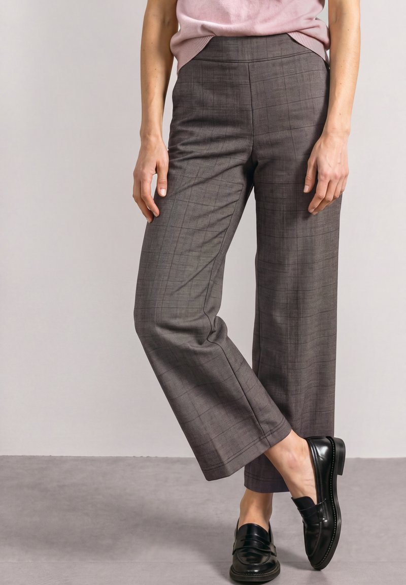 Femme portant un pantalon marron droit à carreaux, un haut rose et des mocassins en cuir noir, debout avec un pied croisé devant l'autre.