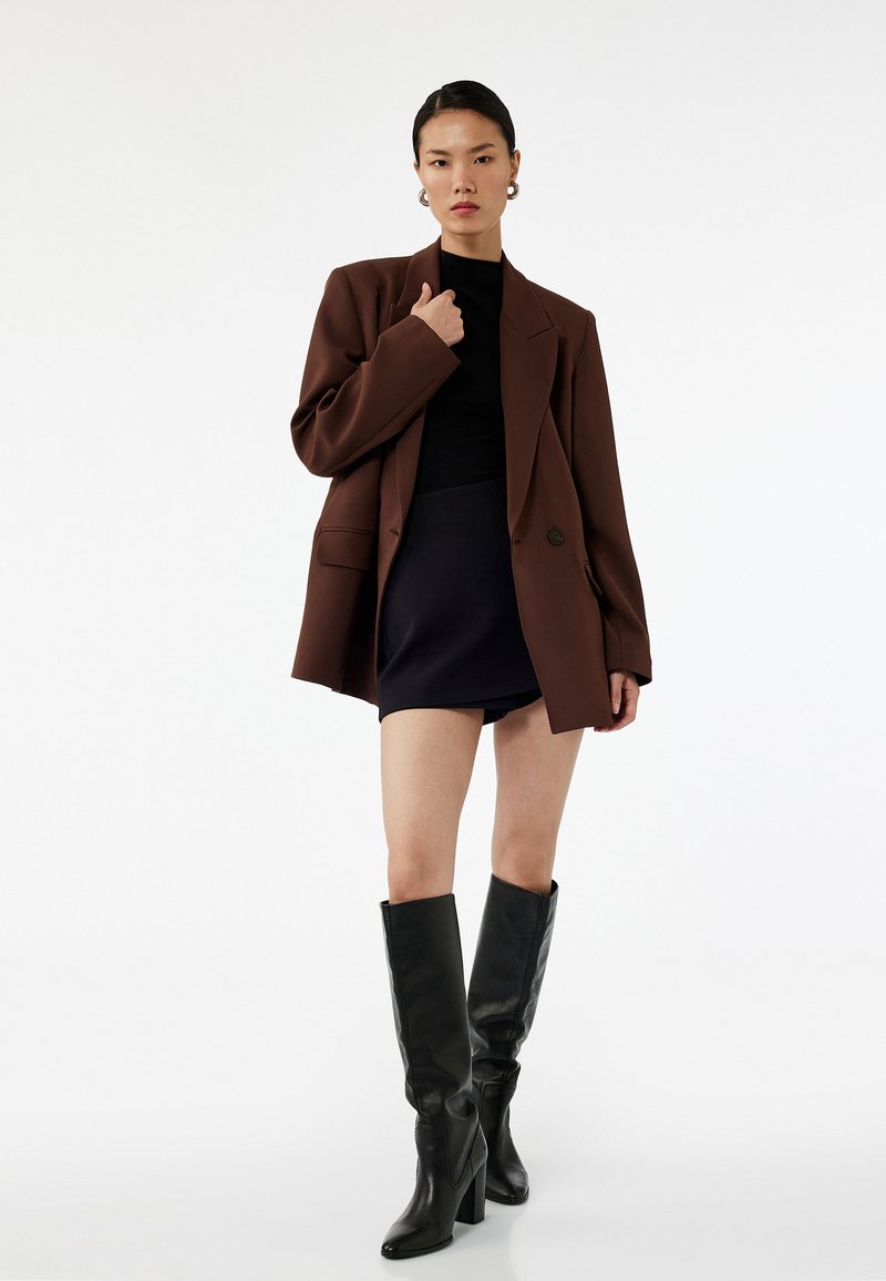 Brun oversized blazer, sort top, sort mini nederdel og knæhøje sorte hælede støvler. Glatte teksturer og strukturerede designelementer.