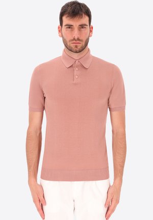 Uomo con capelli corti castani e barba, che indossa una polo aderente rosa polvere a maniche corte e pantaloni bianchi, in piedi davanti a uno sfondo uniforme.