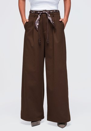 Pantalon marron à jambes larges avec plis à l'avant, poches latérales et ceinture en tissu marron à motif nouée à la taille, porté avec des chaussures grises à bout pointu.