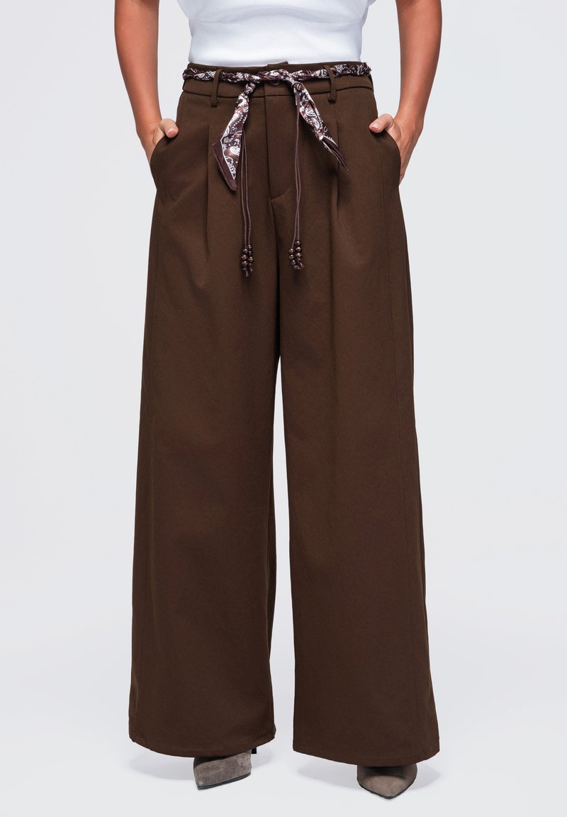 Pantalon marron à jambes larges avec plis à l'avant, poches latérales et ceinture en tissu marron à motif nouée à la taille, porté avec des chaussures grises à bout pointu.