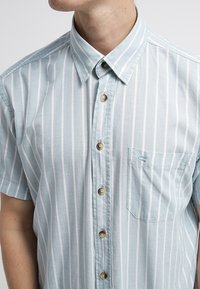 Kurzärmeliges Hemd zum Knöpfen in Hellblau mit weißen vertikalen Streifen, ausgestattet mit einer Tasche und braunen Knöpfen. Weicher Stoff mit lässiger Passform.