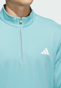 Světlomodrý pulovr s krátkým zipem z měkké látky. Má kovový zip a bílé logo se třemi pruhy na levé straně hrudi. Hladká textura.