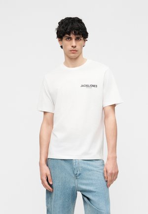 Ung mand med krøllet mørkt hår iført en hvid Jack & Jones Premium t-shirt og lyseblå jeans mod en ensfarvet baggrund.