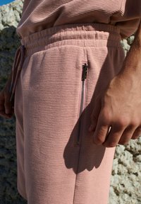 Lyserøde teksturerede shorts med elastisk talje og snørebånd. Har en sidelomme med lynlås. Blødt stof med diskret ribbet mønster.