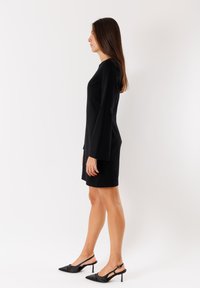 Abito nero con maniche lunghe e una forma aderente, realizzato in tessuto morbido. Abbinato a tacchi neri a punta con cinturini alla caviglia.
