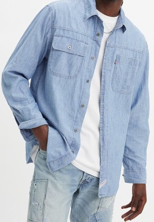 Homme portant une chemise en jean bleu clair boutonnée par-dessus un t-shirt blanc et un jean bleu clair avec des patchs, main dans la poche, sur un fond uni.