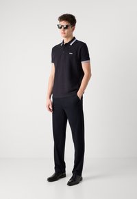 Mornarska polo majica s kontrastnim prugastim ovratnikom, logotipom na prsima, u paru s tamnim hlačama i crnim cipelama. Model nosi sunčane naočale.