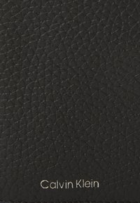 Billetera de cuero negro con textura de guijarros. Presenta un sutil logo en plata en la parte inferior. Diseño rectangular compacto.