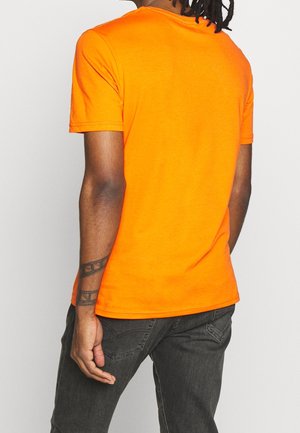 Orange crew neck t-shirt lavet af bomuld. Korte ærmer, tætsiddende design, ensfarvet og minimal detaljering på ryggen. Båret med mørkegrå bukser.