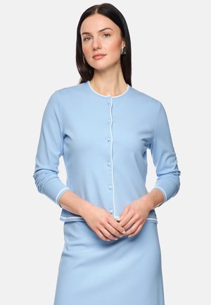 Woman with long dark hair wearing a light blue buttoned cardigan and matching skirt, posing with hands clasped in front.