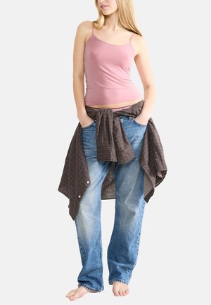 Jasnoróżowy top na ramiączkach, luźne niebieskie jeansy i brązowa koszula w kratę związana w talii. Modelka stoi boso z rękami w kieszeniach.