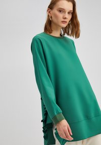 Maglietta a maniche lunghe verde smeraldo con scollatura tonda, dotata di dettagli con volant lungo il lato e polsini a costine. Tessuto morbido e leggero.