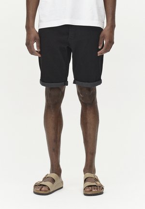 Shorts in denim nero con orli risvoltati, indossati con sandali beige. I pantaloni hanno un taglio classico, mentre i sandali presentano un design senza chiusura.