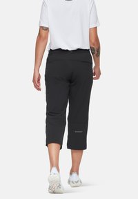 Μαύρο cropped παντελόνι φτιαγμένο από ελαστικό υλικό, με τσέπη με φερμουάρ στο πλάι και διακριτική σήμανση κατά μήκος του ποδιού. Φοριέται με λευκά αθλητικά παπούτσια.