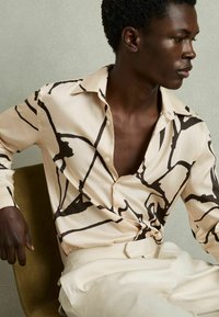 Camisa beige claro con líneas abstractas negras audaces, con un cuello clásico y botones, combinada con pantalones color crema.