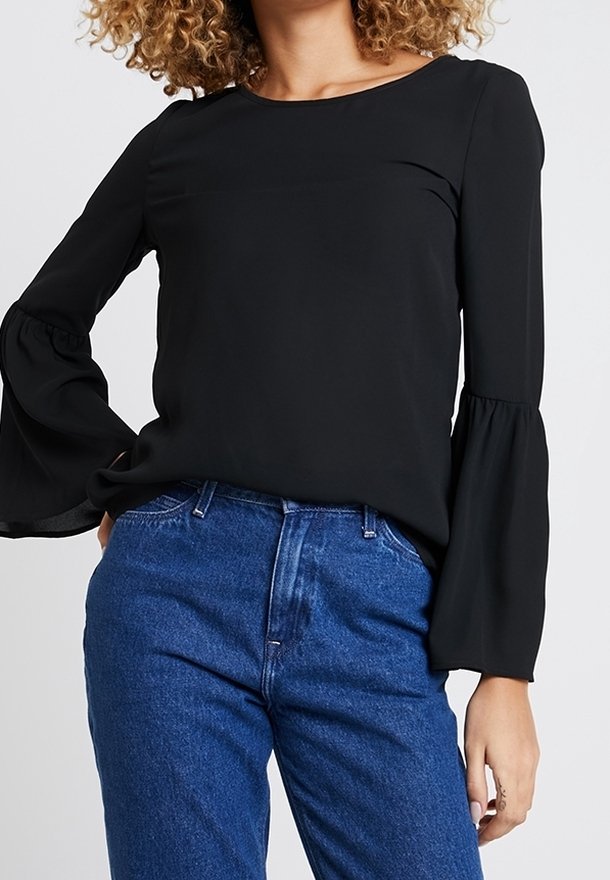 Femme portant un chemisier noir à manches longues évasées et un jean bleu taille haute, debout avec une main sur la hanche.