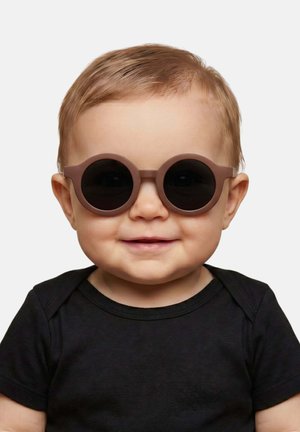Kleinkind trägt runde, dunkle Sonnenbrille und schwarzes Hemd, lächelnd vor einem einfarbigen hellen Hintergrund.