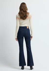 Kremowy sweter prążkowany z otwartym splotem na ramionach, w połączeniu z ciemnoniebieskimi dzwonami z teksturowanym wzorem i brązową skórzaną naszywką z tyłu.