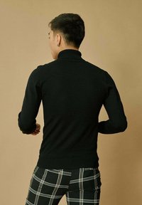 Pull à col roulé noir avec un design ajusté, col et poignets côtelés, associé à un pantalon à motifs plaid. Le fond est d'un beige uni.