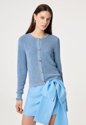 Vrouw die een lichtblauwe gebreide cardigan met zilveren knopen draagt, gecombineerd met een lichtblauwe rok met een grote strik in de taille.