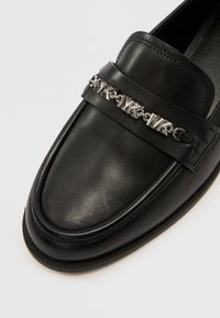 Μαύρο μοκασίνι από δέρμα με στρογγυλεμένη μύτη, διακοσμημένο με ασημένια αλυσίδα στο εμπρός μέρος και εμφανείς ραφές γύρω από τις άκρες.