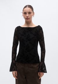 Črna majica z dolgimi rokavi z čipkasto prekrivalo z motivi cvetličja, čolnčkim izrezom in zvončastimi rokavi. Ponaša se z izčrpanimi cvetličnimi vzorci in prosojno teksturo.