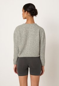 Moterys su tamsiais plaukais, susegtais kuodu, dėvi šviesiai pilką pūkuotą megztinį ir aptemptus tamsiai pilkus šortus, žiūrėdama į nugarą, priešais paprastą foną.