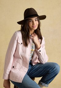 Jeune femme portant un chapeau marron à large bord, une veste rose, un pull à motifs et un jean bleu, assise avec un genou relevé sur un fond beige.