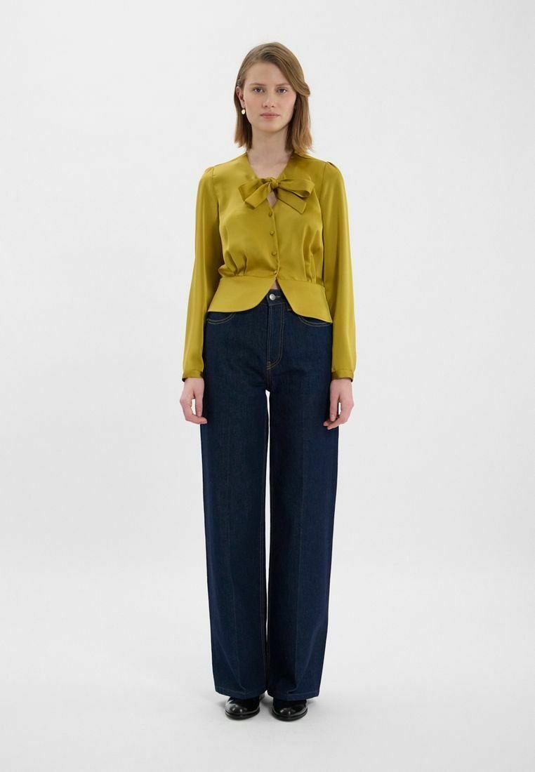 Blouse en satin jaune avec un grand nœud au col, devant boutonnière et ourlet structuré, associée à un pantalon large en denim foncé taille haute.