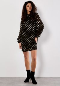 Schwarzes Kleid mit goldenen Punkten, langen Ärmeln und lockerer Passform. Gestylt mit schwarzen Stiefeletten. Glattes Material mit leichter Textur.