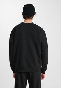 Persona che indossa un berretto in maglia nero, felpa nera e pantaloni neri, in piedi con le spalle rivolte verso un fondo chiaro e semplice.