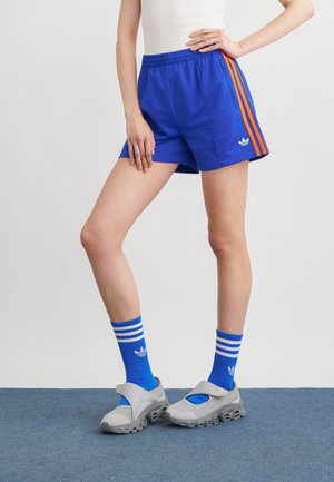 Persoon draagt blauwe sportshorts met oranje strepen en bijpassende blauwe Adidas sokken, gecombineerd met grijze sportieve sneakers, staand op een blauw tapijt.