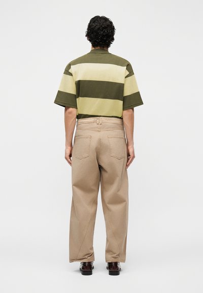 Άνδρας που στέκεται με την πλάτη στην κάμερα, φορώντας πράσινο και μπεζ πουκάμισο με φαρδιές ρίγες και κοντά μανίκια, μπεζ παντελόνι και σκούρα παπούτσια σε λευκό φόντο.