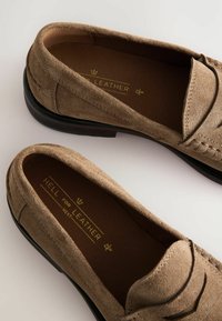 Mocasines de ante marrón con una suela de goma negra suave, que cuentan con detalles de costura y una marca en dorado en la plantilla.