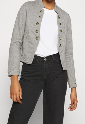 Kobieta w szarej krótkiej kurtce z złotymi guzikami, białej koszulce i czarnych jeansach z wysokim stanem, stojąca z rękami w luźnej pozycji.