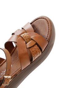 Sandalia de cuero marrón con acentos tejidos, tiras cruzadas y plantilla texturizada. Incluye una ligera suela de cuña y correa ajustable en el tobillo.