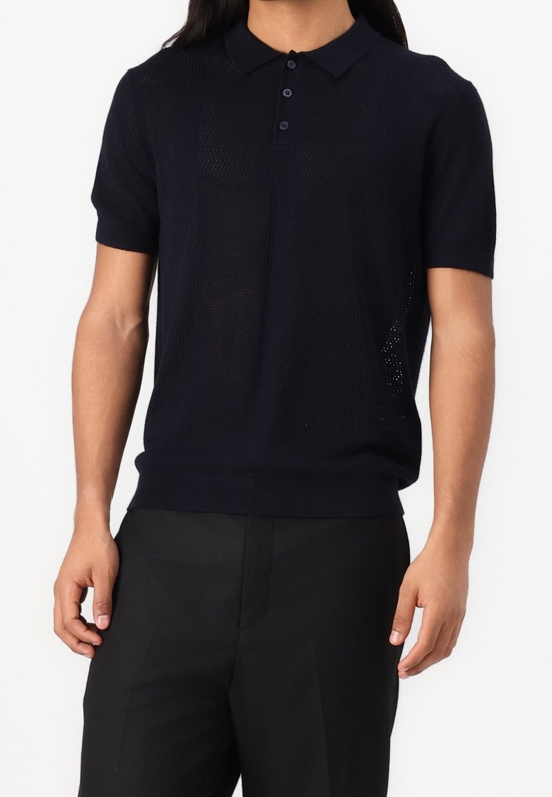 Homme portant un polo à manches courtes en maille bleu marine foncé avec une patte de boutonnage à trois boutons et un pantalon noir sur un fond uni.