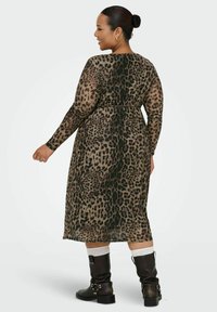 Donna sorridente, che indossa un vestito midi a maniche lunghe con stampa leopardata, calzini bianchi e stivaletti neri in pelle, in piedi contro uno sfondo semplice.
