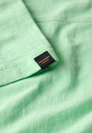 Tissu en coton vert clair à texture douce, plié, avec une étiquette noire embossée de "SUPERDRY PREMIUM" en orange.