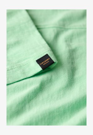 Lichtgroene katoenen stof met een zachte textuur, dubbelgevouwen, met een zwart label waarop in oranje 'SUPERDRY PREMIUM' gedrukt staat.