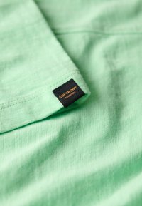 Tela verde menta con un acabado texturizado, que presenta una etiqueta negra con "Superdry" en letra naranja en un borde doblado.