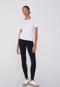 T-shirt blanc à manches courtes associé à un legging noir. Le modèle porte des baskets blanches avec des semelles en caoutchouc, sur fond uni.