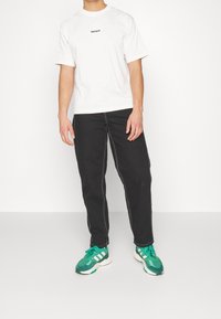 T-shirt en coton blanc avec "WASTEWARE" imprimé en noir, assorti à un pantalon noir avec des coutures contrastées et des chaussures de sport vertes.