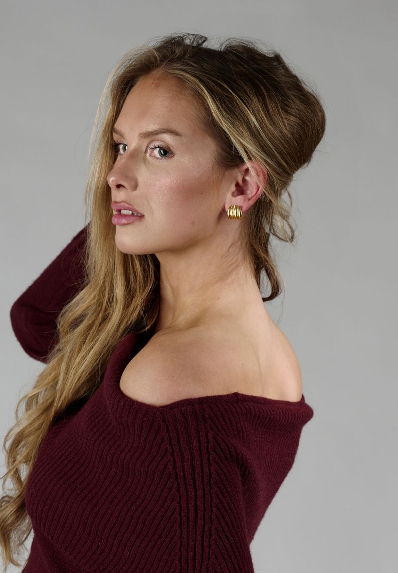 Burgundy off-shoulder ribbet sweater med tekstureret strik, der har en bred halsudskæring og lange ærmer. Gyldne hoops øreringe tilføjer kontrast.