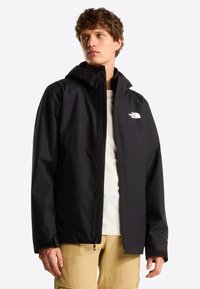 Ung mand iført en sort North Face-jakke over en hvid skjorte og beige bukser, stående mod en ensfarvet baggrund og kigger let mod venstre.