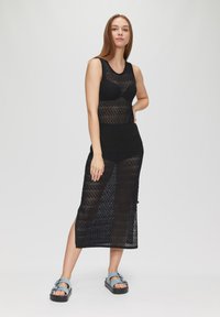 Das schwarze Strickkleid zeichnet sich durch ein tailliertes Oberteil und einen langen, seitlich geschlitzten Rock mit einem strukturierten Diamantmuster aus. Kombiniert mit blauen Plateausandalen.