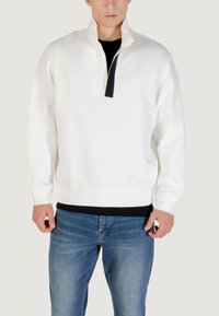 Sudadera blanca con medio cierre en el cuello y detalles en negro, puños y dobladillo acanalados; combinada con jeans de mezclilla azules.